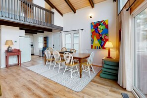 A dining area with a wooden table and white chairs, a colorful abstract painting on the wall, and a balcony with a railing.