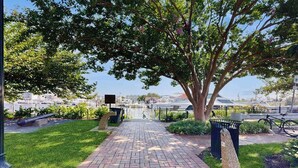 A paved pathway leading to a park area with a large tree, a trash bin, and a bicycle. - Town of Lewes condo steps from the park w/Tennis, Pickleball & Basketball (Lewes)