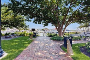 A paved pathway leading to a park area with a large tree, a trash bin, and a bicycle. - Town of Lewes condo steps from the park w/Tennis, Pickleball & Basketball (Lewes)