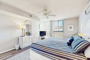 Ein Schlafzimmer mit einem Bett, einem Deckenventilator, einem Schminktisch, einem Fernseher und einem Fenster mit Jalousien.