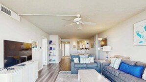 Ein modernes Wohnzimmer mit Flachbildfernseher, Deckenventilator, Sofa, Couchtisch und einer Küche im Hintergrund.