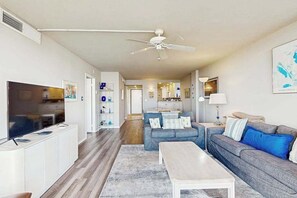 Ein modernes Wohnzimmer mit Flachbildfernseher, Deckenventilator, Sofa, Couchtisch und einer Küche im Hintergrund.