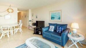 A living room with a blue sofa, a dining table with chairs, a TV, and a lamp. - Sea Colony 7th floor condo with ocean veiw, pool, basketball court &  gym (Bethany Beach)