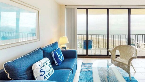 A room with a sofa, a chair, a glass table, and a view of the sea through large windows. - Sea Colony 7th floor condo with ocean veiw, pool, basketball court &  gym (Bethany Beach)