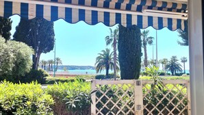 Une vue à travers une clôture en treillis, avec un auvent rayé, des palmiers et une étendue d’eau au loin.