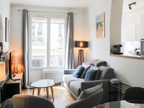A living room with a sofa, a coffee machine, a lamp, and a window with curtains. - Bright Apartment with Montmartre View - 38m² (Paris)