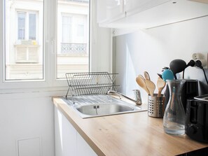 A kitchen with a sink, a dish rack, wooden utensils, a glass pitcher, and a coffee maker. - Bright Apartment with Montmartre View - 38m² (Paris)