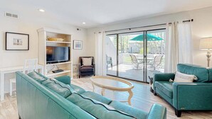 A living room with a green sofa, a TV, a coffee table, and a sliding glass door leading to an outdoor patio. - Sea Colony Tennis 1st floor condo w/ tennis court, basketball court, and pool (Bethany Beach)
