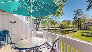 A deck with a table and chairs, a green umbrella, and a view of a pond and trees. - Sea Colony Tennis 1st floor condo w/ tennis court, basketball court, and pool (Bethany Beach)