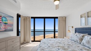 A bedroom with a large bed, a ceiling fan, a nightstand, a lamp, a dresser, and a view of the beach. - Golden Sands 1018 by Seaside Vacations & Sales (Ocean City)
