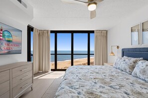 Ein Schlafzimmer mit einem großen Bett, einem Deckenventilator, einem Nachttisch, einer Lampe, einer Kommode und Blick auf den Strand.