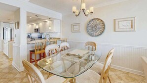 A dining area with a glass table, white chairs, a chandelier, and a kitchen in the background. - Sea Colony condo in oceanside complex with washer/dryer, shared hot tub & pool (Bethany Beach)