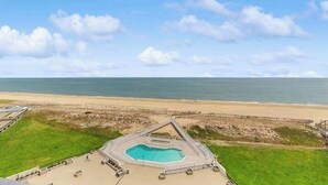 A swimming pool with a geometric design, surrounded by a paved deck and a grassy area, with a beach and ocean in the background. - Sea Colony condo in oceanside complex with washer/dryer, shared hot tub & pool (Bethany Beach)