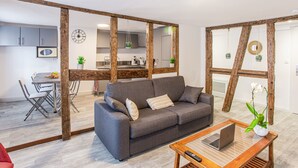 Una sala moderna con un sofá gris, vigas de madera, un comedor con mesa y sillas, y una cocina visible a través de una puerta abierta.