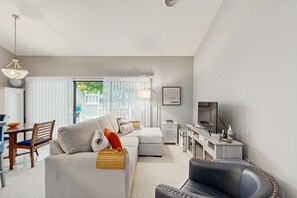 A modern living room with a sofa, a dining table, a TV, and a lamp. - Sea Colony Woodland retreat with pool & hot tub, gym, central AC, WiFi, deck (Bethany Beach)
