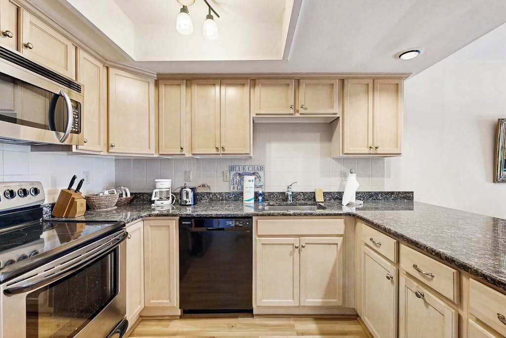 Una cocina moderna con armarios de madera clara, encimera de granito, microondas integrado, lavavajillas y cafetera.