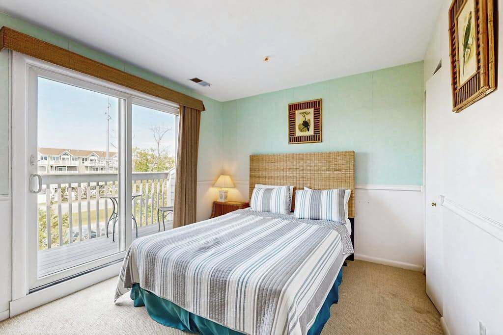 Ein Schlafzimmer mit einer großen Schiebeglaswand, einem Bett mit gestreifter Bettwäsche, einem Rattan-Kopfschild und einem gerahmten Bild an der Wand.