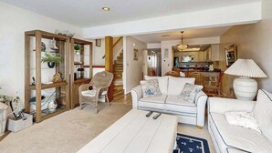 A spacious living area with a white sofa, a coffee table, and a bookshelf. There is a staircase leading to another level, and a kitchen area is visible in the background. - Indian Harbor Villas home close to the beach with private washer/dryer & WiFi (Bethany Beach)