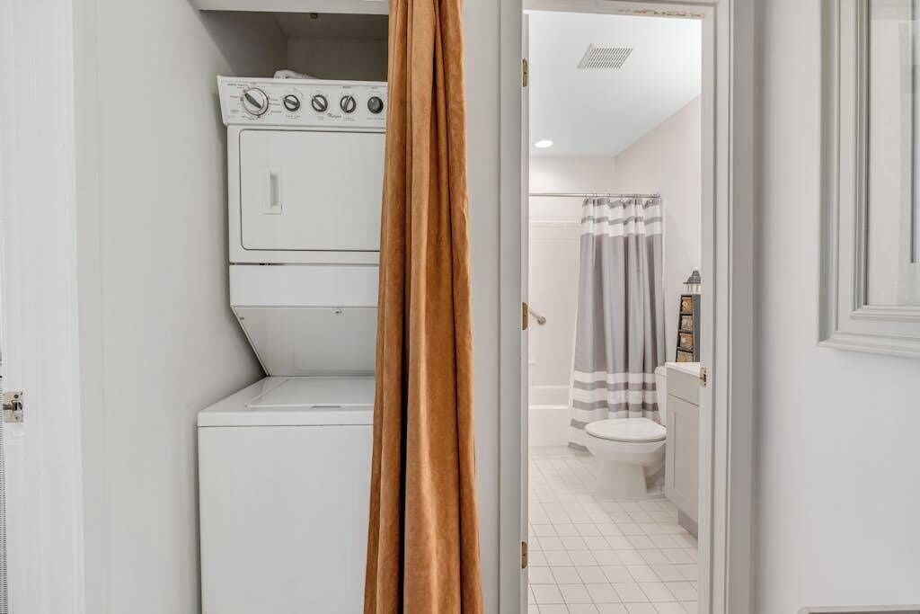 Une buanderie équipée d’un lave-linge et d’un sèche-linge, une salle de bain avec des toilettes et un rideau de douche, et une porte dont un rideau marron recouvre partiellement la vue.