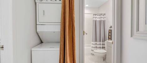 Une buanderie équipée d’un lave-linge et d’un sèche-linge, une salle de bain avec des toilettes et un rideau de douche, et une porte dont un rideau marron recouvre partiellement la vue.