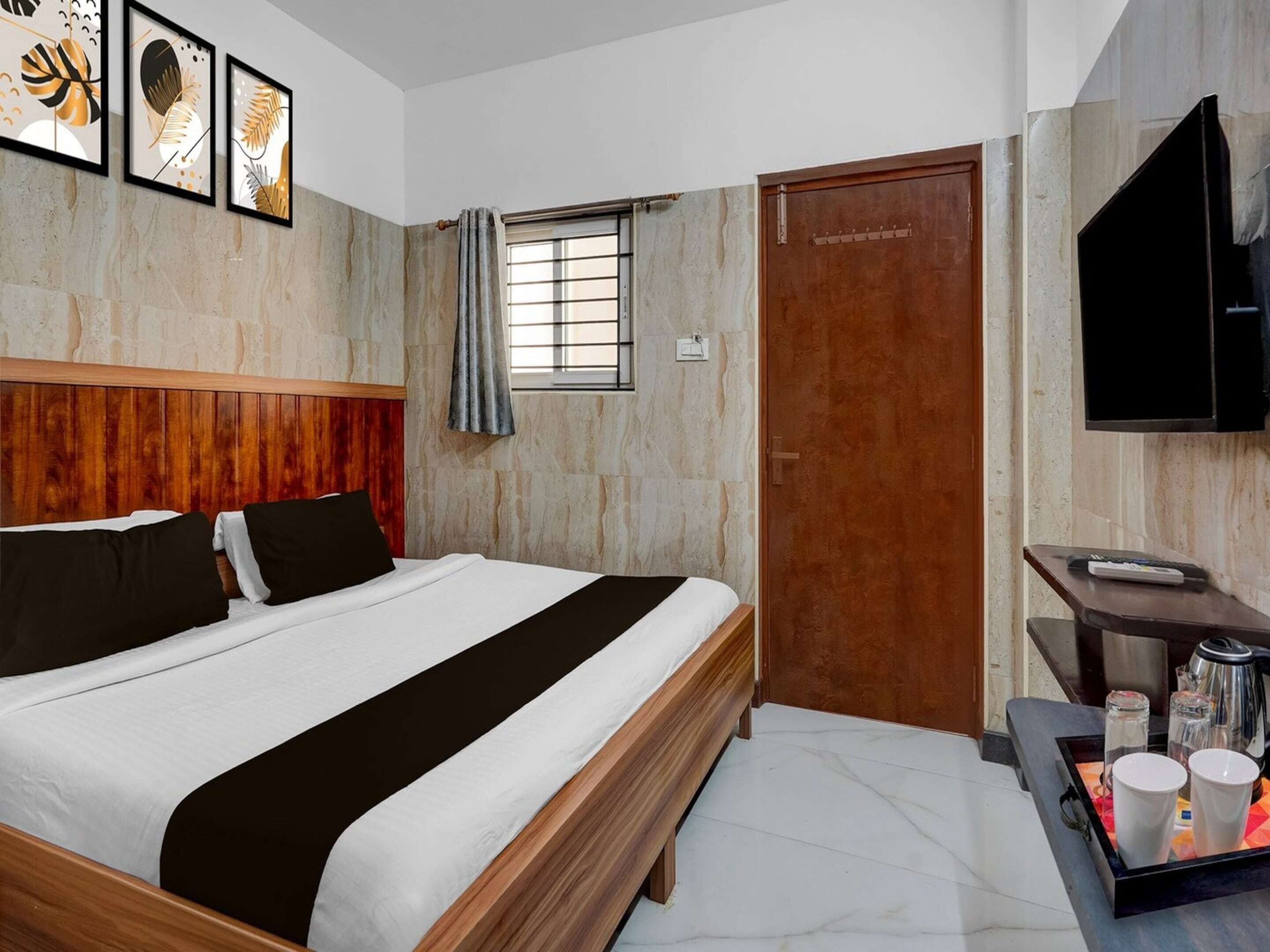 Una habitación de hotel con cabecera de madera, cama con ropa de cama blanca y negra, ventana con cortinas, puerta de madera, televisor de pantalla plana y una estación de café.