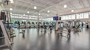 Un gimnasio amplio con diversas máquinas de ejercicio, ventanas grandes y un techo alto.