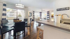 Et moderne køkken med en central ø, et spiseområde med et glasbord, og udsigt til et hus gennem store vinduer.