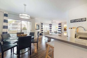 A modern kitchen with a central island, a dining area with a glass table, and a view of a house through large windows. - Sea Colony Waterfront Condo w/Deck, Private Beach, Shared Pools, Hot Tub, Tennis (Bethany Beach)