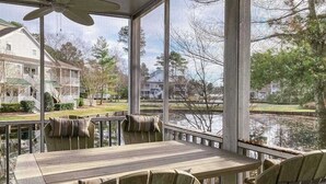 Et overdækket udendørs område med et bord, stole, store vinduer og udsigt til sø og huse.