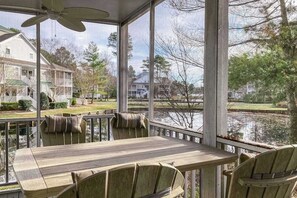 A covered outdoor area with a table and chairs, large windows, and a view of a lake and houses. - Sea Colony Waterfront Condo w/Deck, Private Beach, Shared Pools, Hot Tub, Tennis (Bethany Beach)