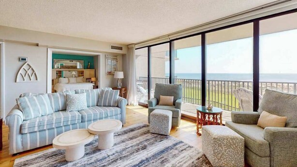 Un salon spacieux avec une grande fenêtre donnant sur la mer, un canapé, deux tables d’appoint et un tapis à motifs.