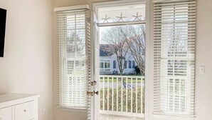 Une chambre à coucher comprenant un lit, une commode et une porte donnant sur un balcon agrémenté de volets blancs, d’où l’on peut admirer une maison et des arbres.