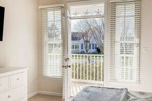 Ein Schlafzimmer mit einem Bett, einer Kommode und einer Tür, die auf einen Balkon mit weißen Fensterläden und Blick auf ein Haus und Bäume führt.
