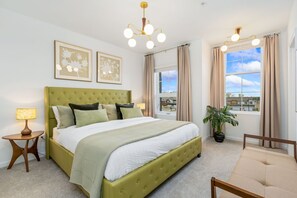 A neatly arranged bedroom with a green upholstered bed, a bedside lamp, a window with curtains, and a view of buildings outside.