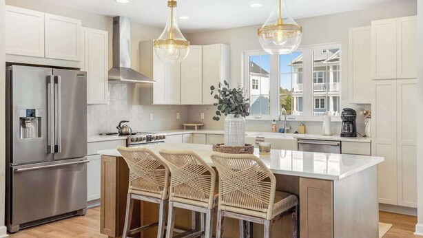 A modern kitchen with a stainless steel refrigerator, a central island with bar stools, pendant lighting, and a view of a house through a window. - Luxe New Home in Tidal Walk - 2 Decks, Home Office, Dogs OK, AC & Shared Pool (Ocean View)