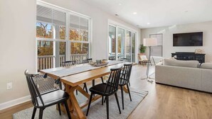 A modern living room with a large window, a dining area with a wooden table and chairs, and a grey sofa. - Luxe New Home in Tidal Walk - 2 Decks, Home Office, Dogs OK, AC & Shared Pool (Ocean View)