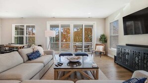 A spacious living room with a sofa, coffee table, and a TV mounted on the wall. - Luxe New Home in Tidal Walk - 2 Decks, Home Office, Dogs OK, AC & Shared Pool (Ocean View)