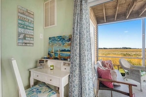 A room with a desk, a chair, a painting, and a view of a field through a window. - Bethany Bay condo with pool, fireplace, screened porch & AC (Ocean View)