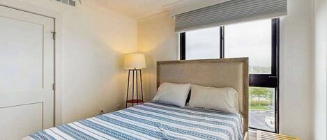 A bedroom with a bed, a nightstand, a lamp, a window, and a view of the outside.