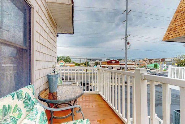 A balcony with a table and chairs, a white railing, and a view of the street and buildings in the background.
