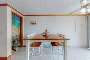 A dining area with a wooden table and white chairs, a ceiling fan, and a door with a view of the outside.