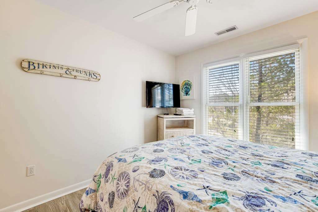 A bedroom with a bed, a television, a window with blinds, and a wall-mounted sign.