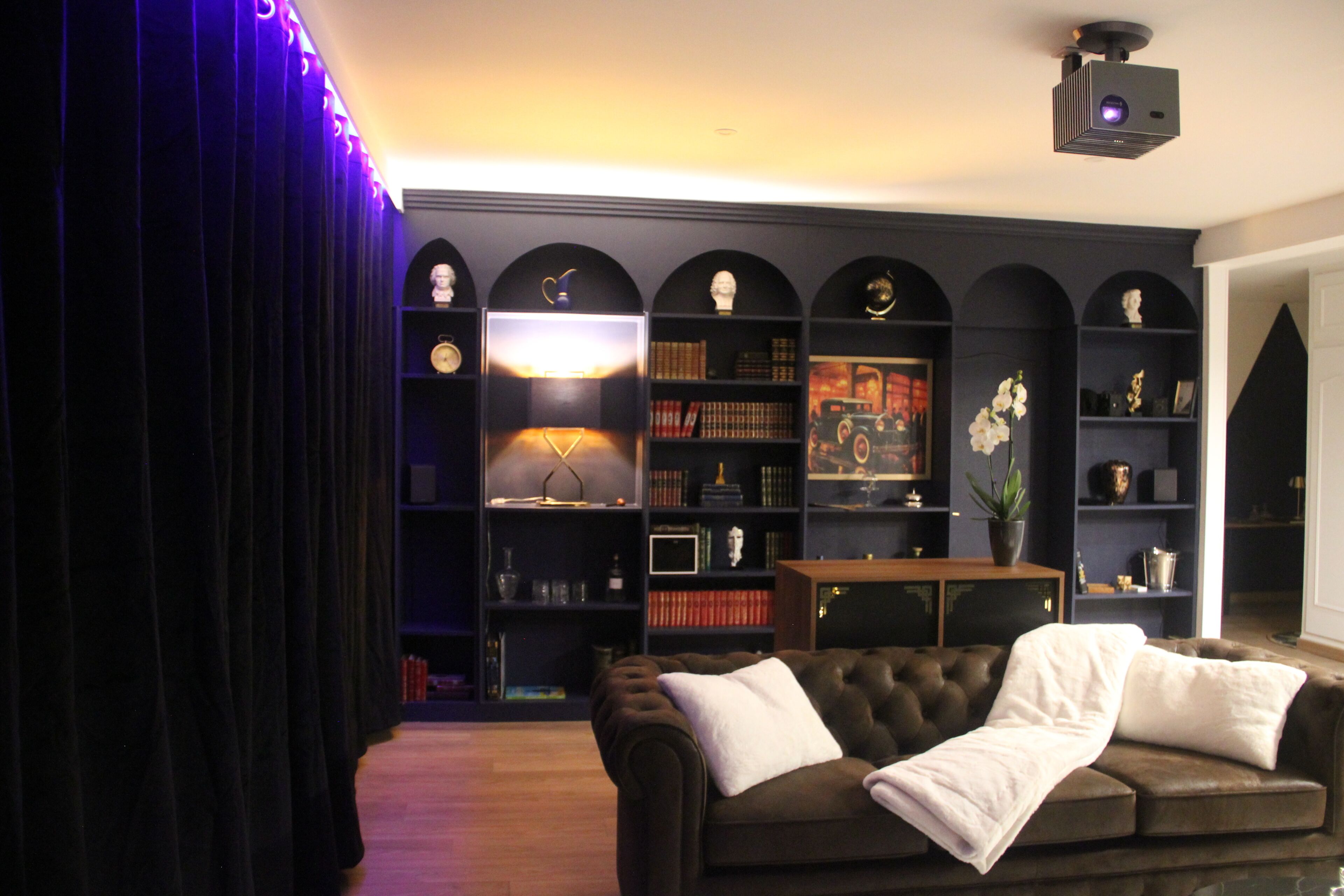 Een kamer met een donkergrijze bank, een boekenplank met diverse spullen, en een aan het plafond bevestigde beamer.