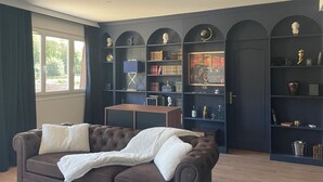 A room with a dark blue arched bookshelf, a brown leather sofa with white cushions, a projector on the ceiling, and a window with curtains. - Le Repaire du Gentleman - Spa, Private Cinema & King-Size Bed • Enclosed Garage (Étretat)