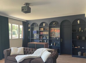 Ein Zimmer mit einem dunkelblauen Bogenregal, einem braunen Ledersofa mit weißen Kissen, einem Projektor an der Decke und einem Fenster mit Vorhängen.