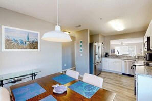 Une cuisine moderne équipée d’appareils électroménagers en acier inoxydable, un coin repas avec une table en bois et des sets de table bleus, et une vue sur la mer par la fenêtre.