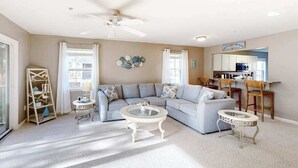 A living room with a sectional sofa, a round coffee table, a small side table, a ceiling fan, and a kitchen area with cabinets and a microwave. - 2nd-floor condo at Bethany Bay with pool, on-site golf, and sports courts (Ocean View)