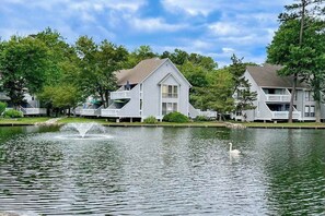A lakeside view of a residential area with white houses, a fountain, and a swan. - Sea Colony Tennis townhouse with pools, tennis, fitness centers, & private beach (Bethany Beach)