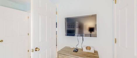 A room with a television mounted on the wall, a wooden dresser with drawers, and white doors with brass handles.