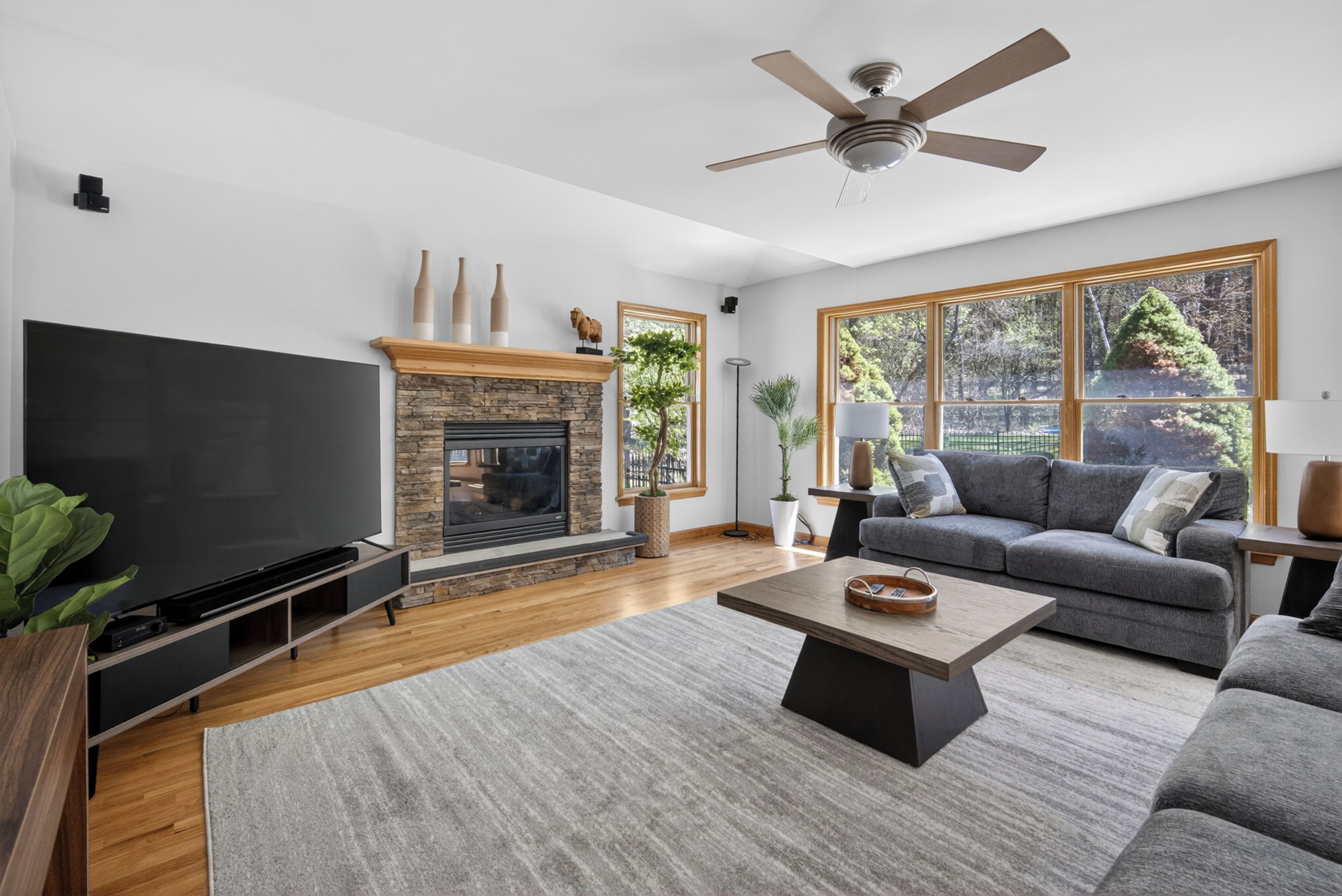 A living room with a flat-screen TV, a fireplace, a ceiling fan, and large windows.